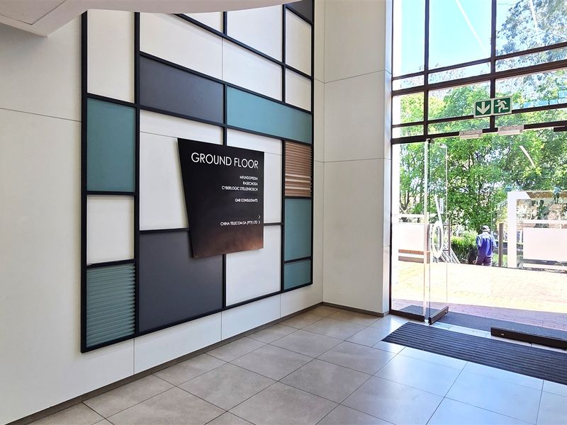 Modern office building entrance lobby featuring high ceilings, large glass doors, tiled flooring, and a contemporary decorative wall panel with directory signage.