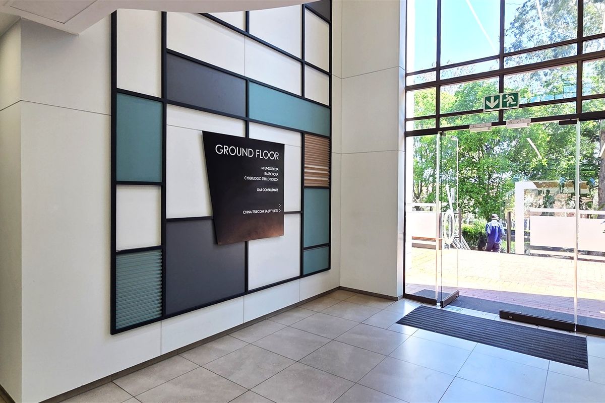 Modern office building entrance lobby featuring high ceilings, large glass doors, tiled flooring, and a contemporary decorative wall panel with directory signage.