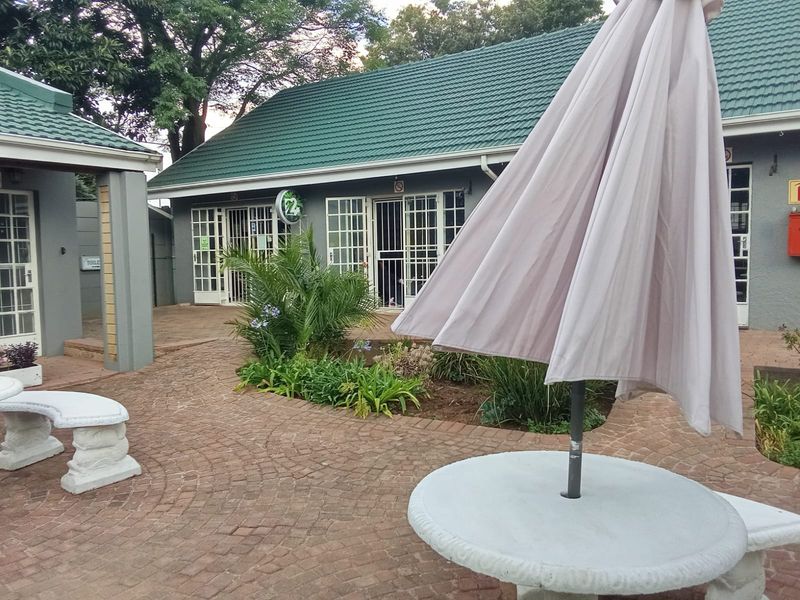 A paved outdoor courtyard area featuring a brick walkway, landscaped garden beds, and access to the main building structure with green tiled roofing.