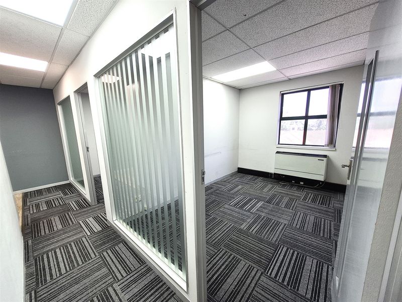 A bright, vacant office space featuring modern carpet tiles, glass partition walls, and integrated HVAC unit, suitable for a professional work environment.