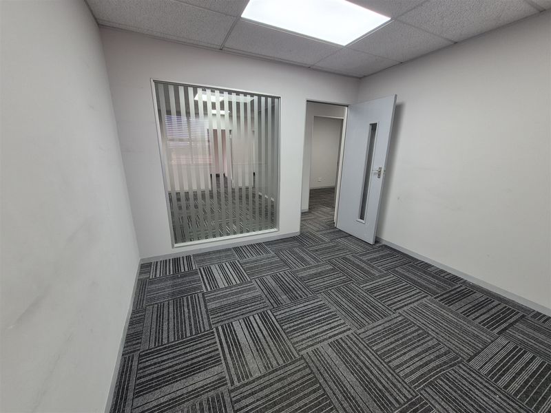 An empty office space featuring carpet tiles, a suspended ceiling with recessed lighting, and an internal glass partition with vertical blinds.
