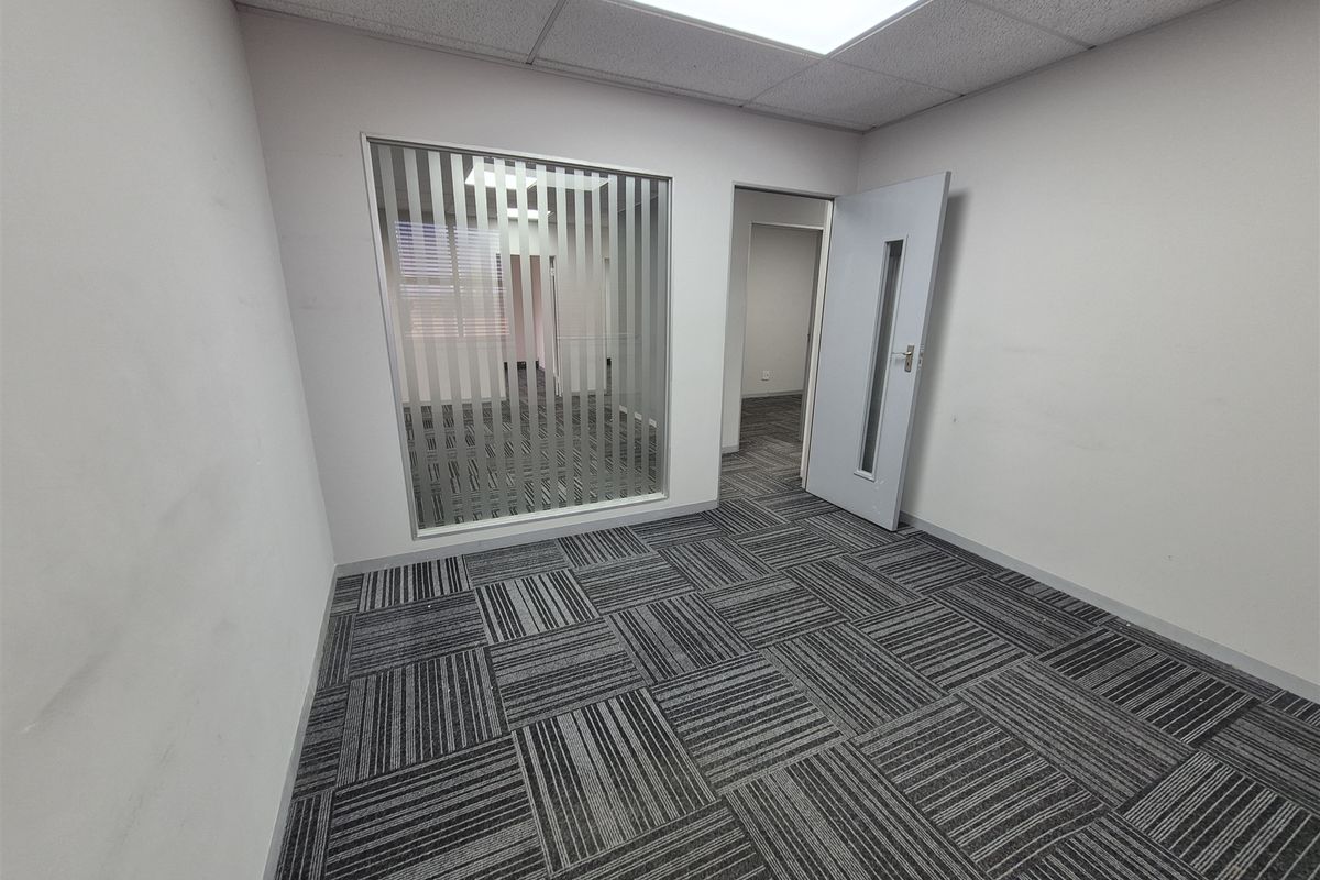 An empty office space featuring carpet tiles, a suspended ceiling with recessed lighting, and an internal glass partition with vertical blinds.