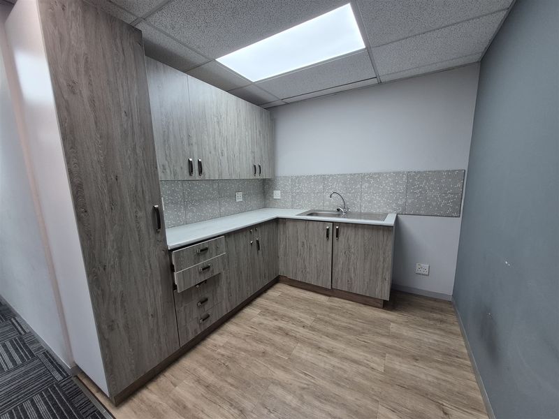 A clean and modern office kitchenette featuring wood-grain cabinetry, a stainless steel sink, tiled backsplash, and durable laminate flooring.