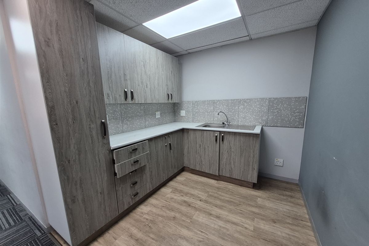 A clean and modern office kitchenette featuring wood-grain cabinetry, a stainless steel sink, tiled backsplash, and durable laminate flooring.