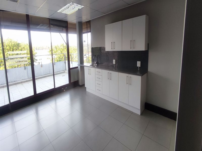 A clean, modern office kitchenette featuring white cabinetry, stainless steel countertops, tiled backsplash, and direct access to a private balcony.