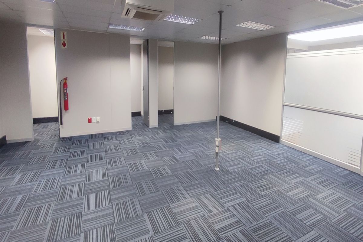An empty, open-plan office space featuring carpet tiles, a suspended ceiling with recessed lighting, a central power pole, and a glass partition wall.