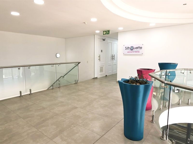A bright and modern office hallway featuring tiled flooring, a glass balustrade overlooking the lower level, and recessed ceiling lighting.