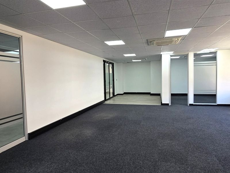 A spacious, empty open-plan office area featuring modern glass partitions, carpeted flooring, and a suspended ceiling with integrated LED lighting.