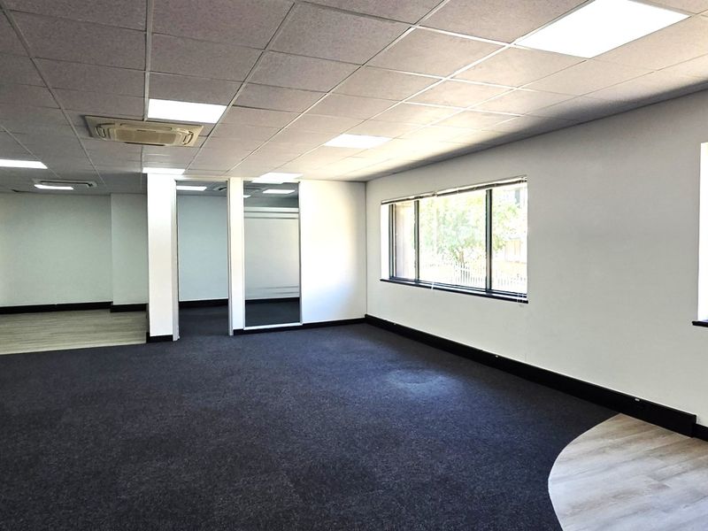 Spacious open-plan office area featuring a mix of carpet and laminate flooring, suspended ceilings with recessed lighting, and large windows providing natural light.