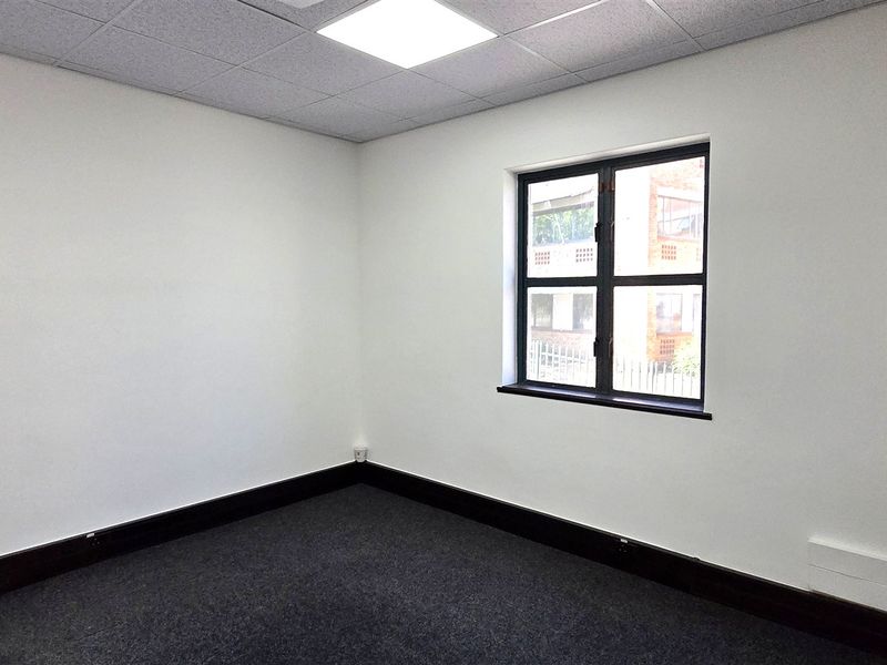 A clean, empty office space featuring neutral walls, dark carpeting, a suspended ceiling with recessed lighting, and a window providing natural light.