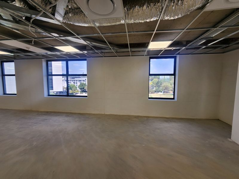 An unfinished open-plan office space featuring exposed ceiling ducting, a suspended grid system, and large windows providing natural light and exterior views.