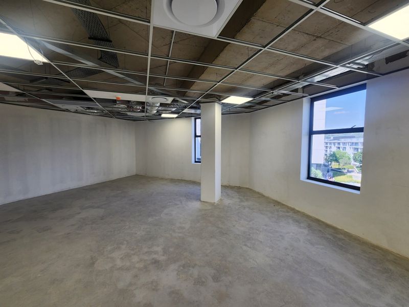 An open-plan office space featuring an unfinished ceiling grid, exposed concrete flooring, and large windows providing natural light and exterior views.