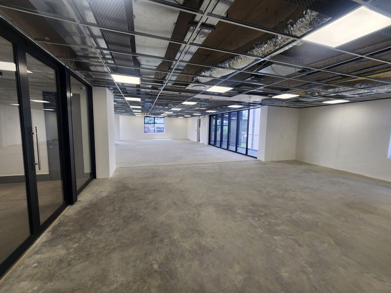An expansive, open-plan office space featuring an industrial-style exposed ceiling grid, recessed lighting, concrete flooring, and floor-to-ceiling glass partitions.