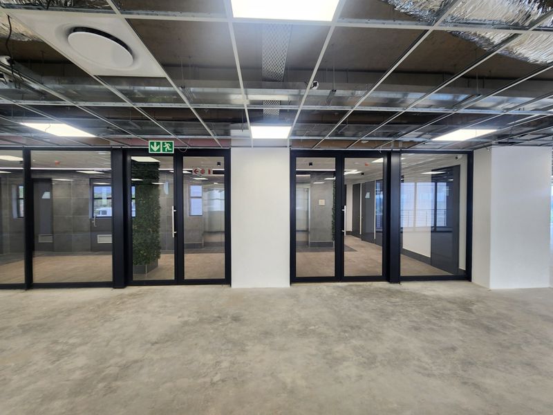 An unfinished office space featuring glass partitions, concrete flooring, and an exposed suspended ceiling grid with integrated lighting and air conditioning.