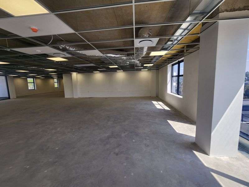 An expansive, open-plan office space currently in a shell condition with exposed ceiling infrastructure, concrete floors, and large windows providing natural light.