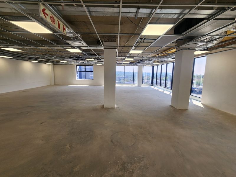 An open-plan office space in shell condition featuring large windows, exposed ceiling grid, and concrete flooring, ready for custom fit-out.