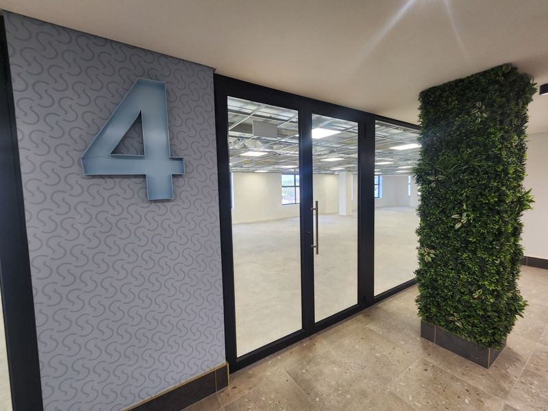 Modern office entrance featuring a glass partition, patterned accent wall, and a decorative artificial green wall, leading into an open-plan workspace with exposed ceiling.