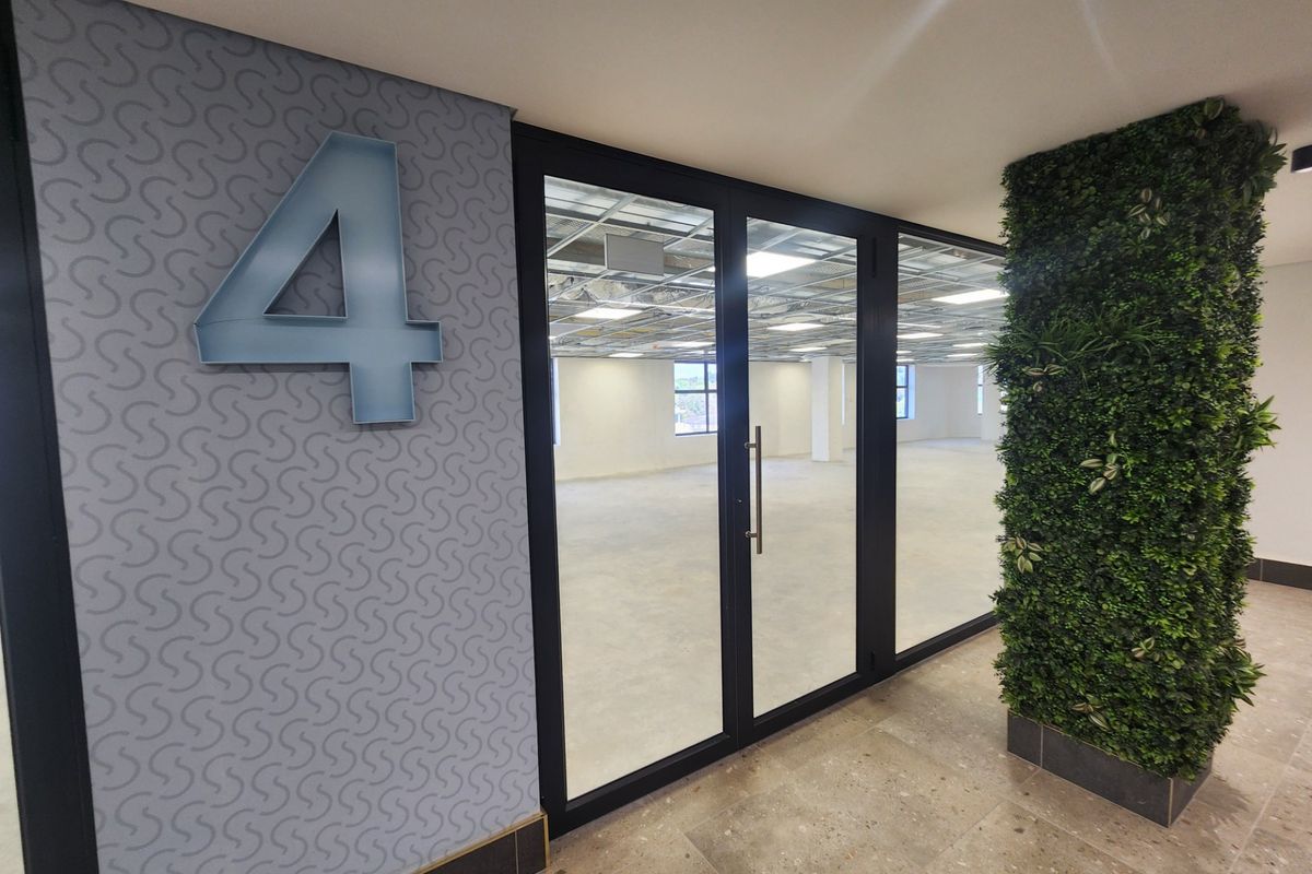 Modern office entrance featuring a glass partition, patterned accent wall, and a decorative artificial green wall, leading into an open-plan workspace with exposed ceiling.