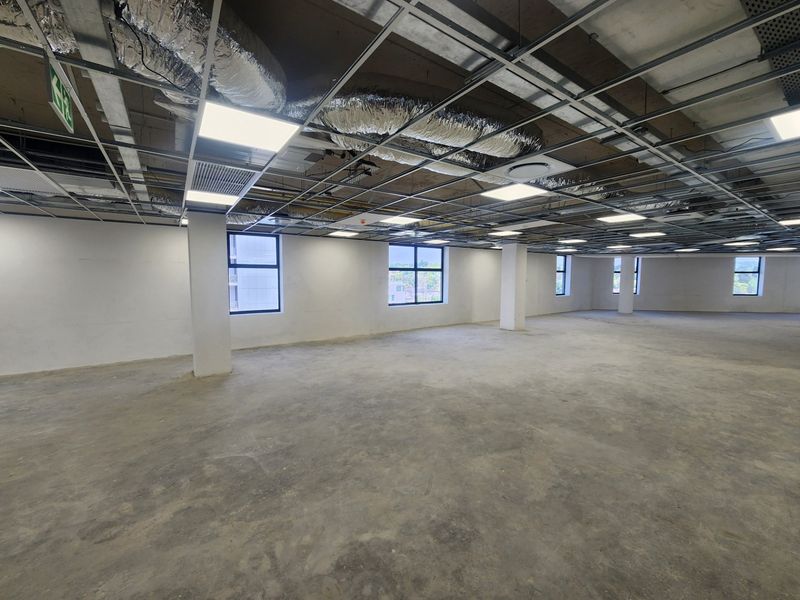 A large, open-plan office space featuring an unfinished ceiling with exposed HVAC ducting, concrete flooring, and ample natural light from multiple windows.