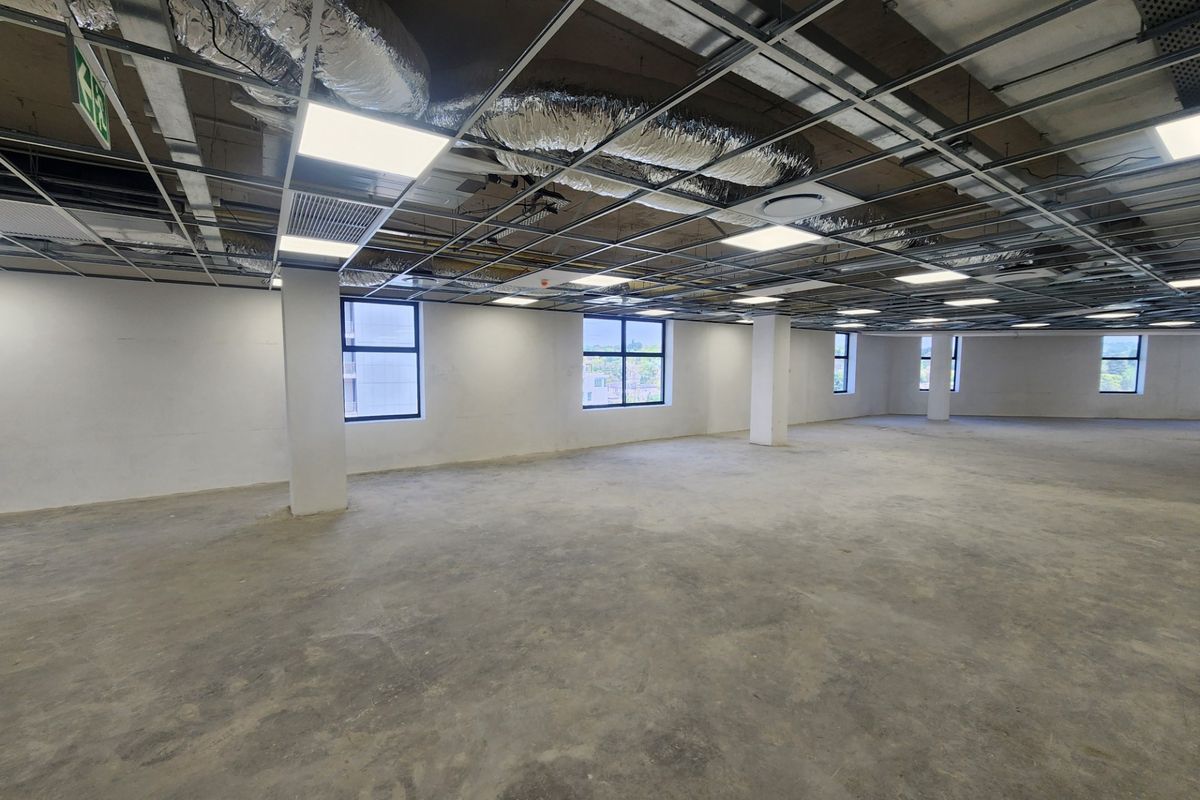 A large, open-plan office space featuring an unfinished ceiling with exposed HVAC ducting, concrete flooring, and ample natural light from multiple windows.