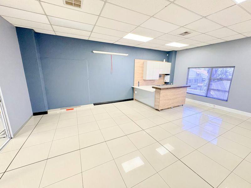 A bright, open-plan office space featuring modern tiled flooring, a suspended ceiling with recessed lighting, and a compact kitchenette area.