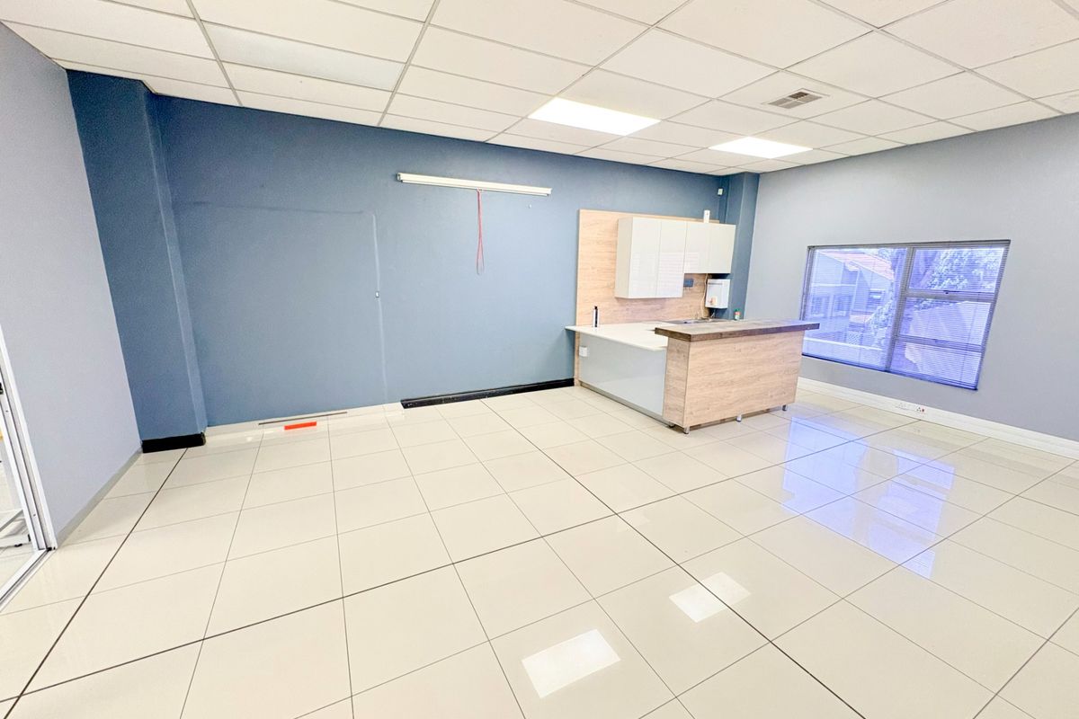 A bright, open-plan office space featuring modern tiled flooring, a suspended ceiling with recessed lighting, and a compact kitchenette area.