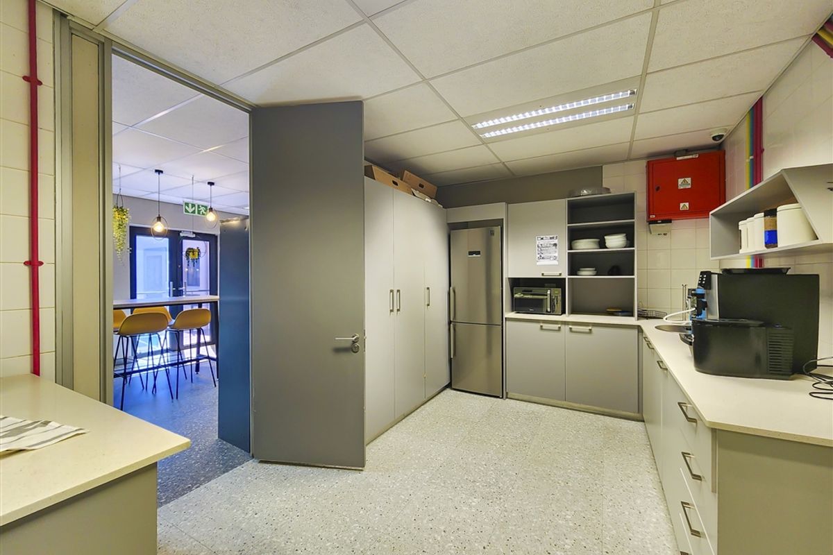 A functional office kitchenette featuring built-in grey cabinetry, laminate countertops, a stainless steel refrigerator, and a drop ceiling with integrated lighting.