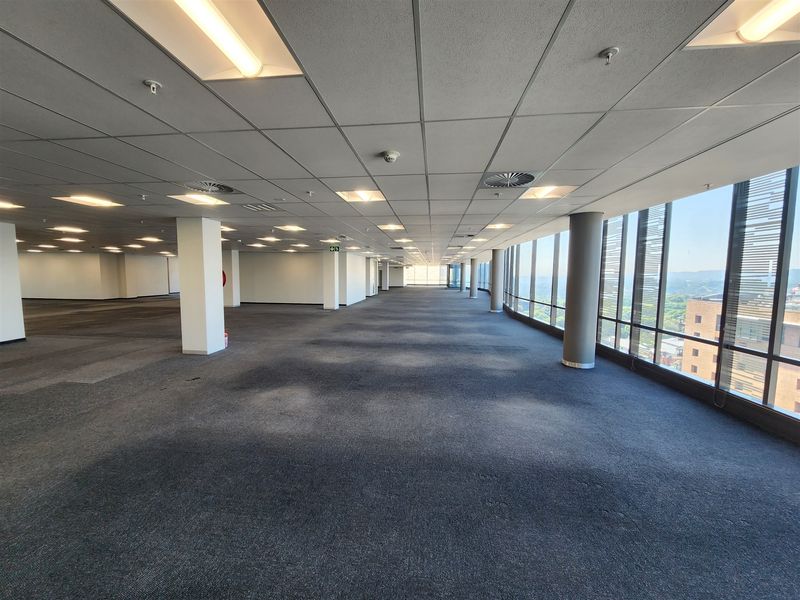 Spacious open-plan office area featuring large windows with panoramic views, suspended ceilings, and carpeted flooring in a well-lit, professional environment.