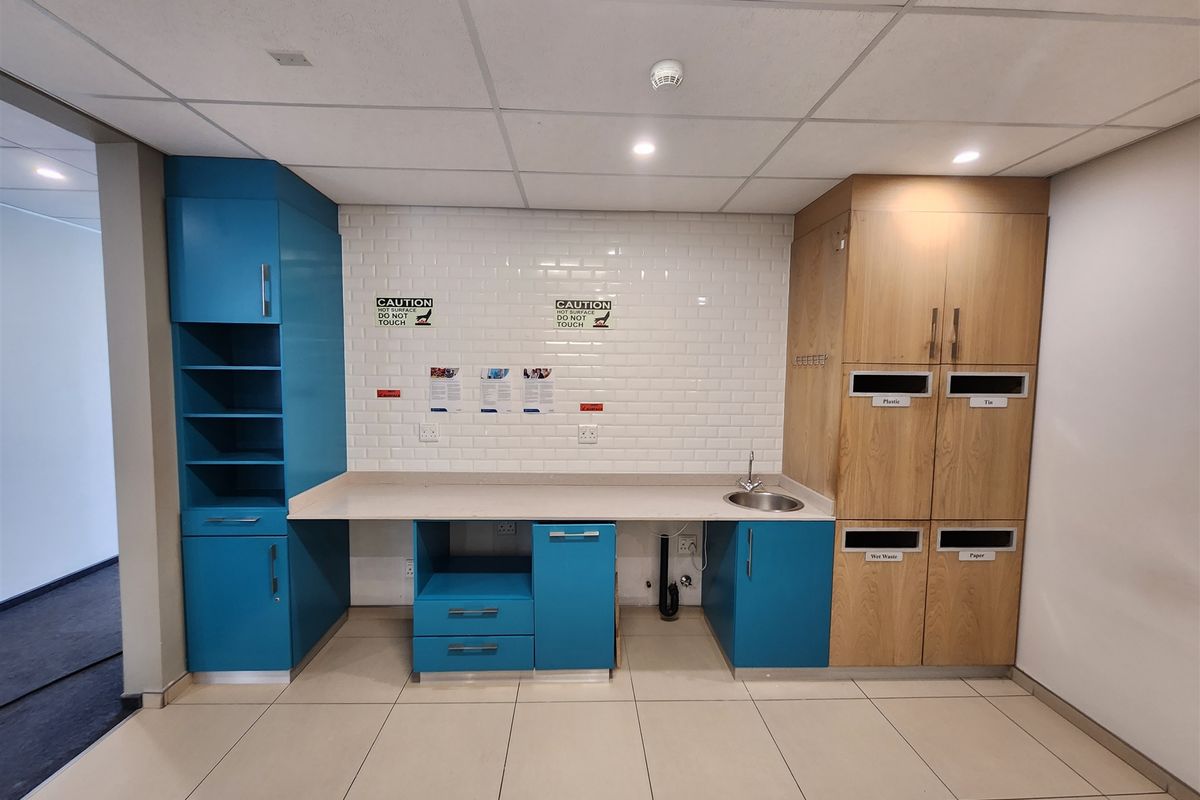 A clean, modern office kitchenette featuring blue and wood-tone cabinetry, a subway tile backsplash, integrated recycling bins, and a small sink.