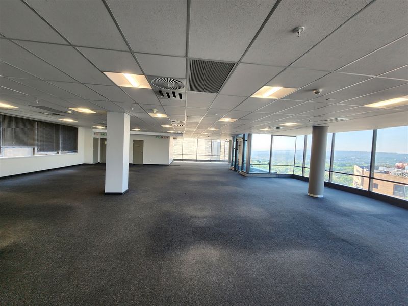 A spacious, open-plan office area featuring large floor-to-ceiling windows with panoramic views, suspended ceilings, and neutral carpeting throughout.