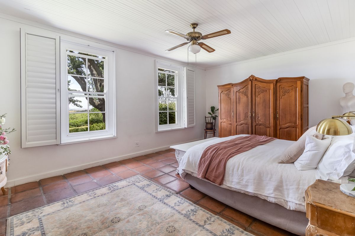 A spacious bedroom featuring traditional terracotta floor tiles, white panelled ceiling, and large windows with fitted shutters offering garden views.