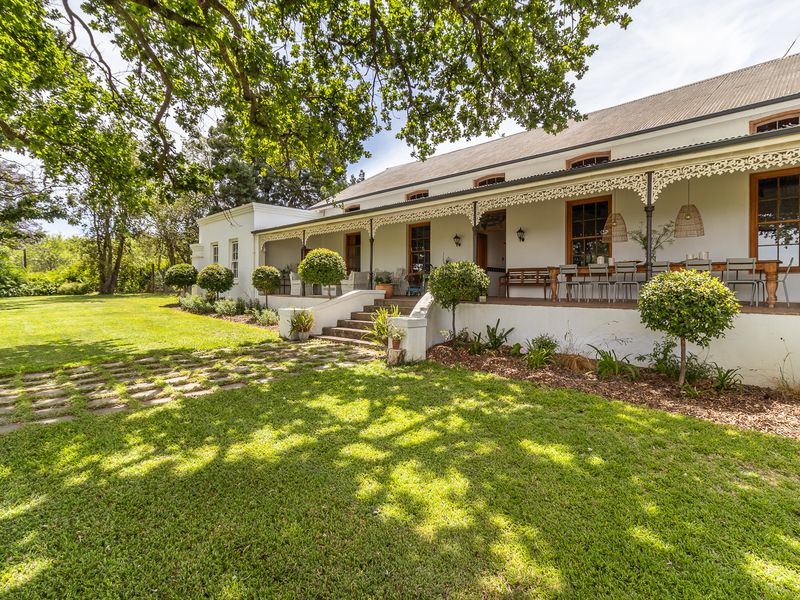 A charming farmhouse exterior featuring a wide covered veranda with decorative ironwork, overlooking a lush green lawn and mature garden landscape.