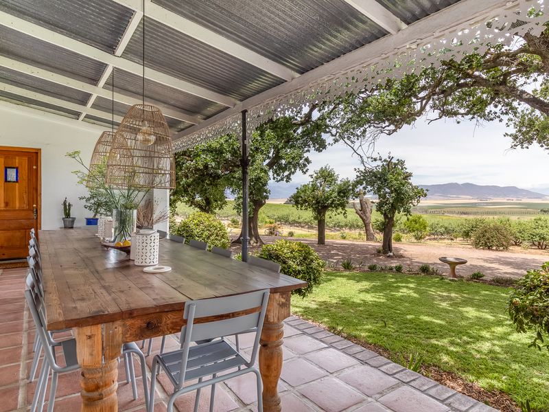 A covered patio featuring terracotta tiled flooring, decorative timber fretwork, and a corrugated metal roof, overlooking a scenic farm landscape with mature trees.
