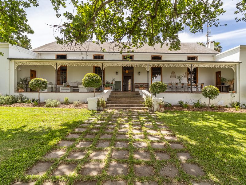 A charming farmhouse exterior featuring a wide covered veranda with ornate ironwork, stone steps, and a paved walkway leading through a lush garden.