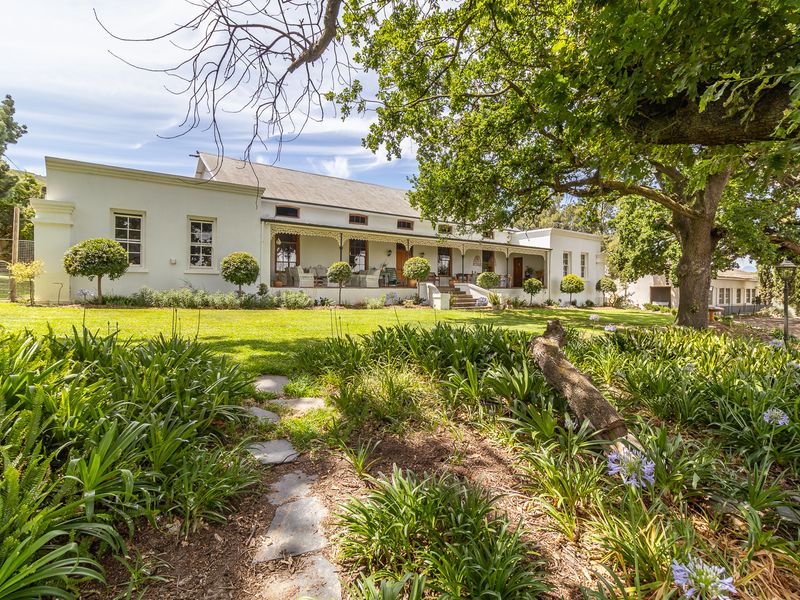 A charming farmhouse exterior featuring a covered veranda, manicured lawn, and established garden beds set against a picturesque rural landscape.