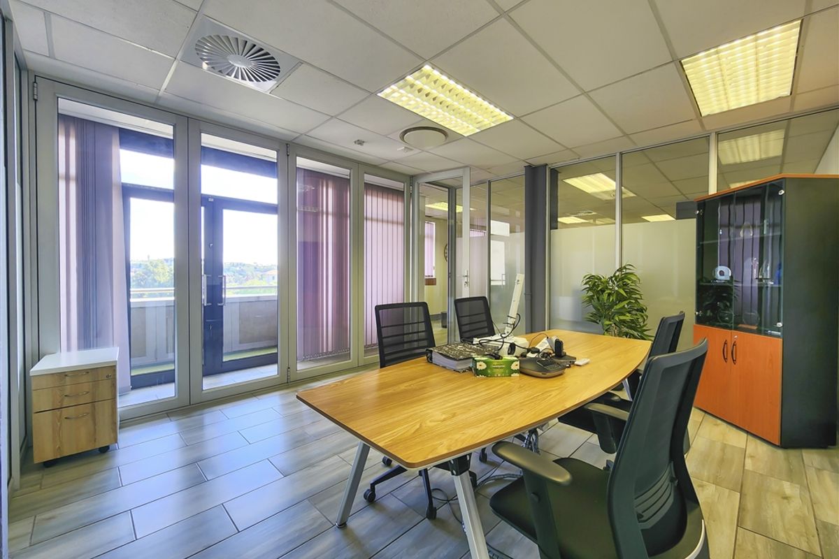 A professional office space featuring glass partition walls, suspended ceilings with recessed lighting, and direct access to an outdoor balcony area.