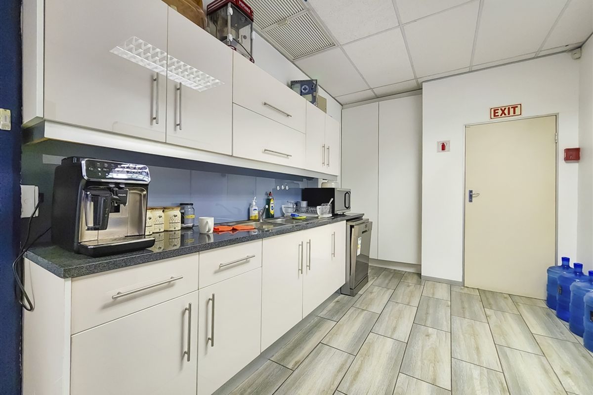 A functional office kitchenette featuring white cabinetry, a dark laminate countertop, stainless steel sink, and durable wood-look tiled flooring.