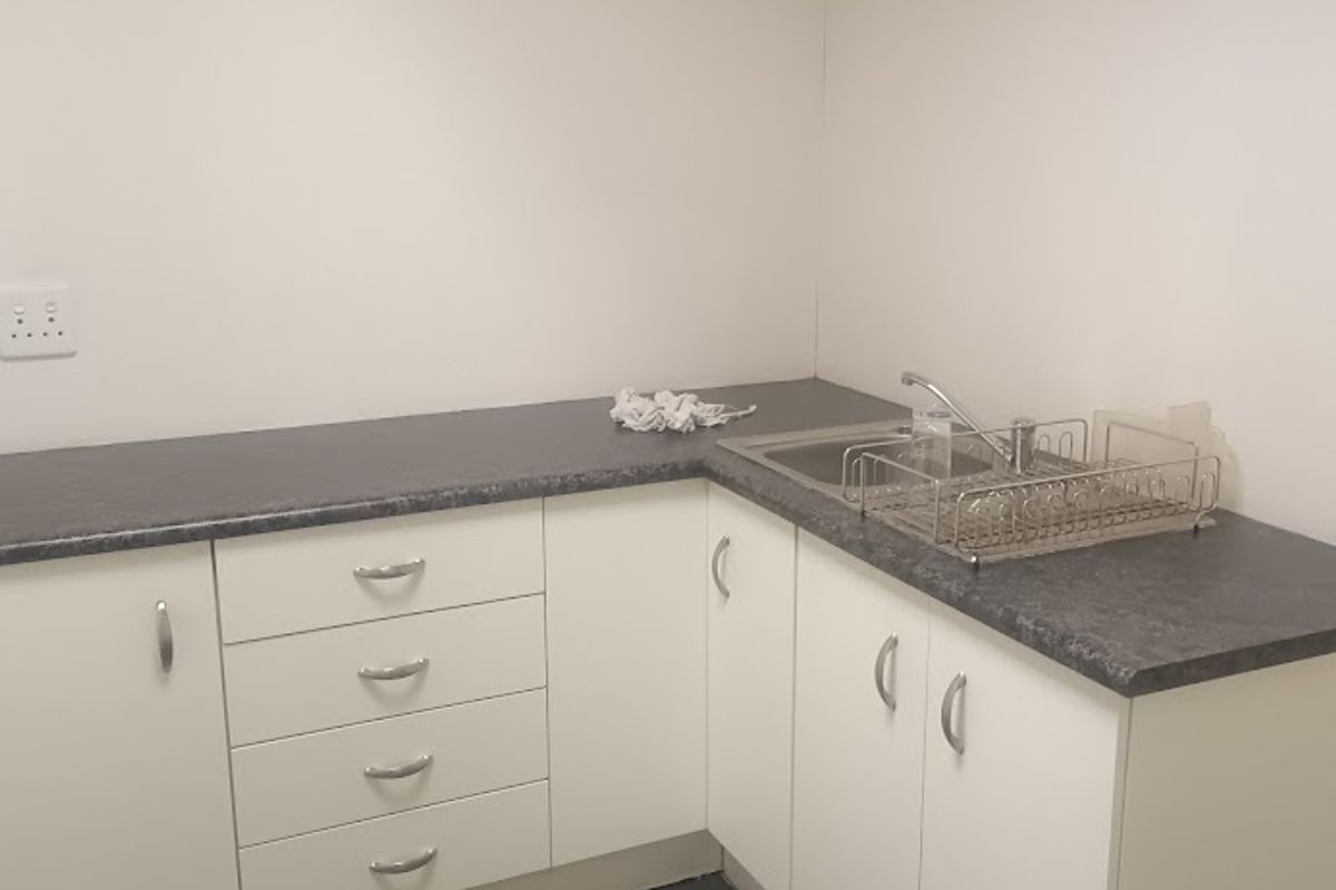 A functional office kitchenette featuring white cabinetry, dark laminate countertops, a stainless steel sink, and dark tiled flooring.