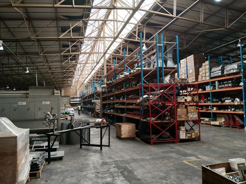 Spacious industrial warehouse featuring high-clearance steel truss roofing, natural skylight panels, durable concrete flooring, and extensive integrated racking systems for storage.