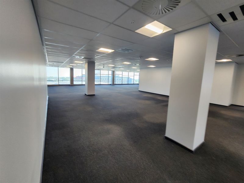 A spacious, open-plan office area featuring large windows, carpeted flooring, and a suspended ceiling with integrated lighting and air conditioning.