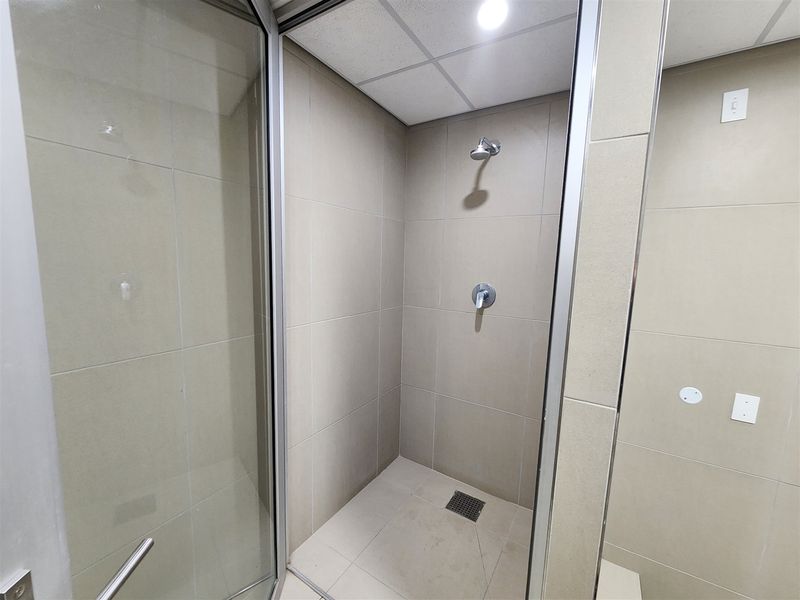 A clean, modern shower stall featuring neutral wall and floor tiling, a glass enclosure, and a drop ceiling with integrated lighting.