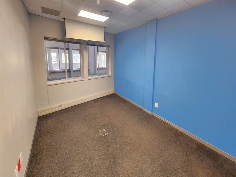 A vacant office space featuring carpeted flooring, a suspended ceiling with recessed lighting, a retractable projector screen, and integrated floor power outlets.