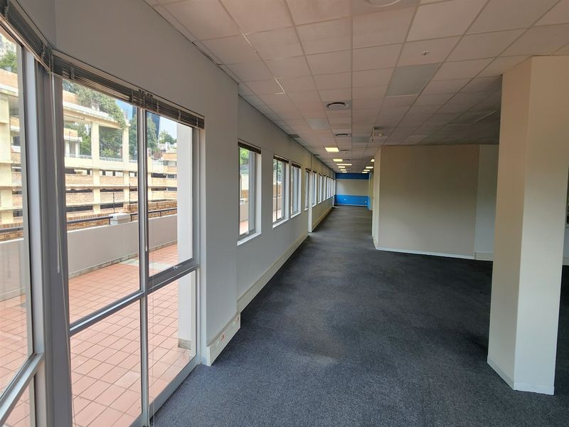 An empty, open-plan office space featuring a suspended ceiling, recessed lighting, carpeted flooring, and large windows with access to an outdoor balcony area.