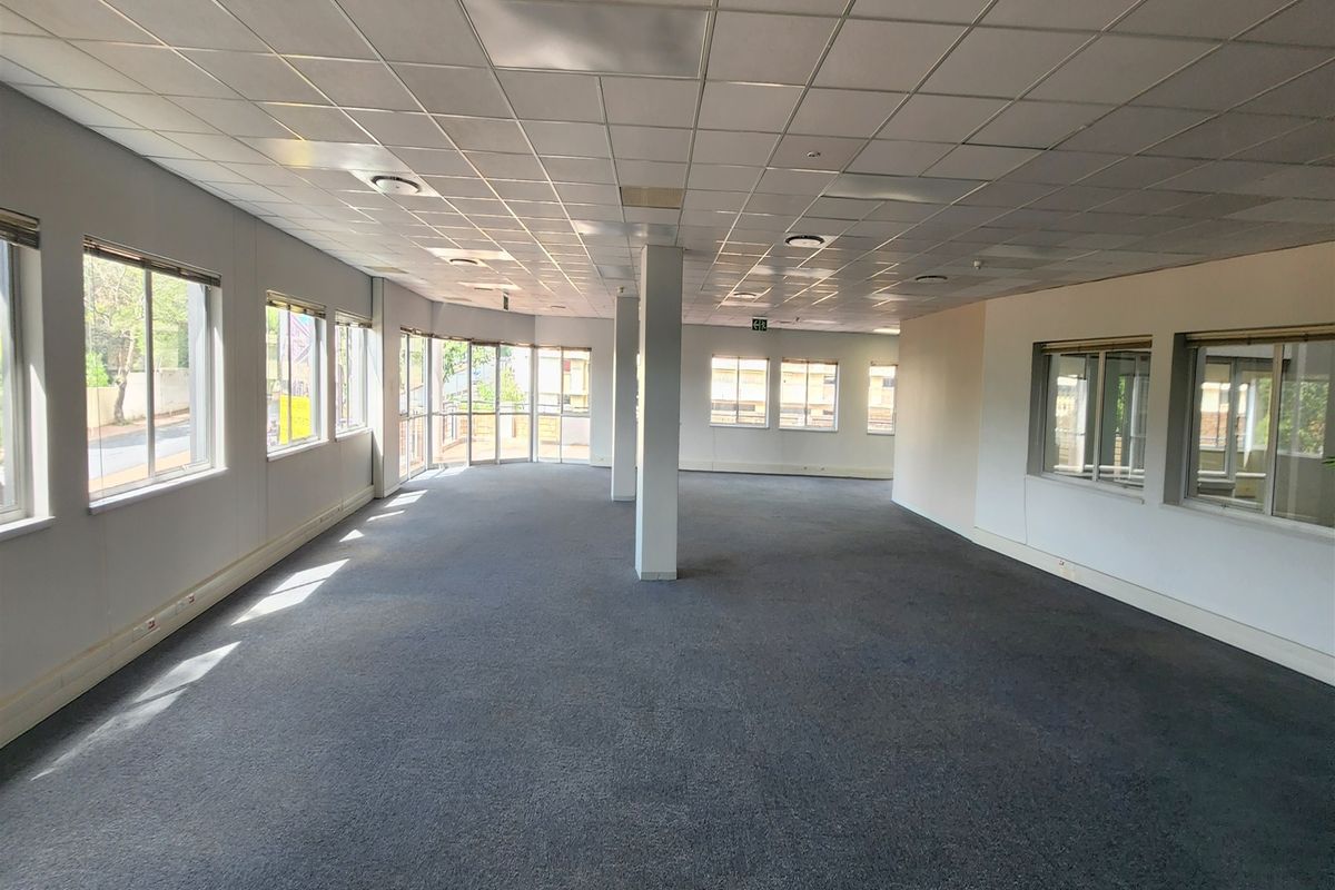 Spacious, open-plan office area featuring a suspended ceiling, large windows providing natural light, and carpeted flooring throughout.