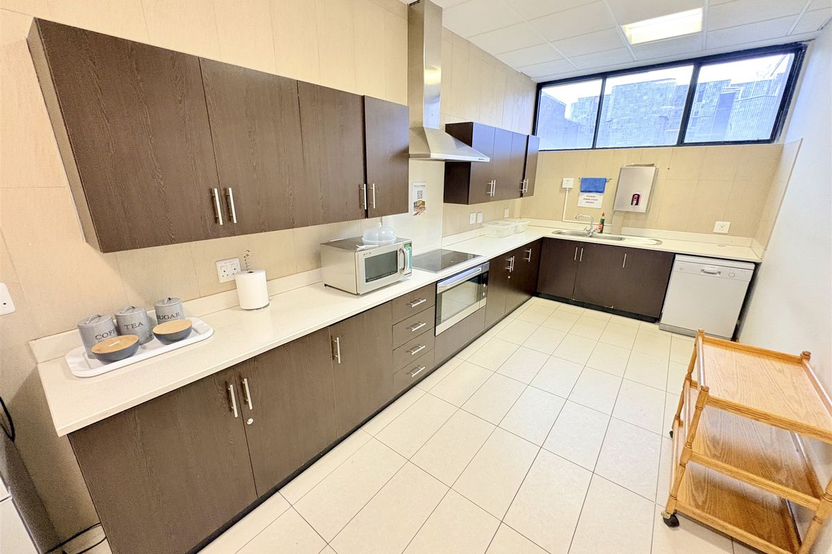 A functional office kitchen featuring dark wood cabinetry, integrated appliances including an oven and extractor fan, and durable tiled flooring.