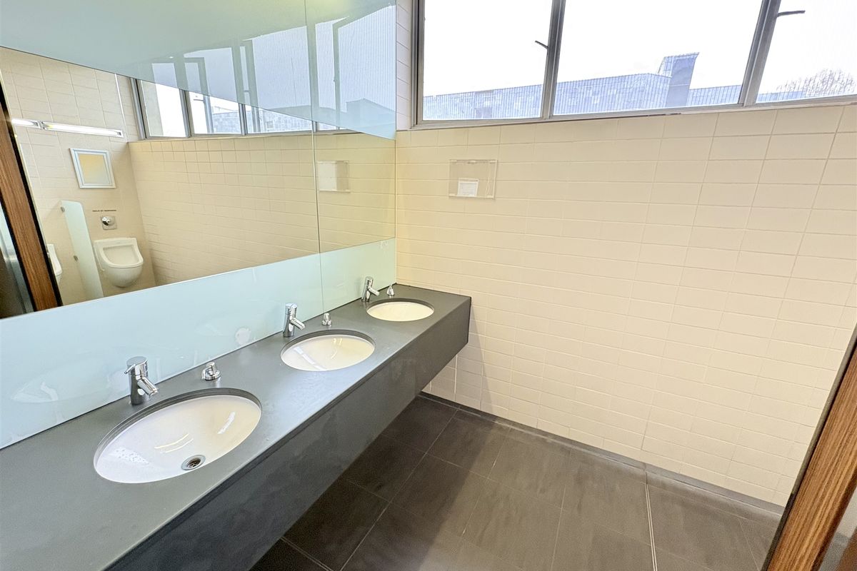 A clean, modern office bathroom featuring a triple vanity basin with integrated sinks, large wall-mounted mirror, and tiled finishes.