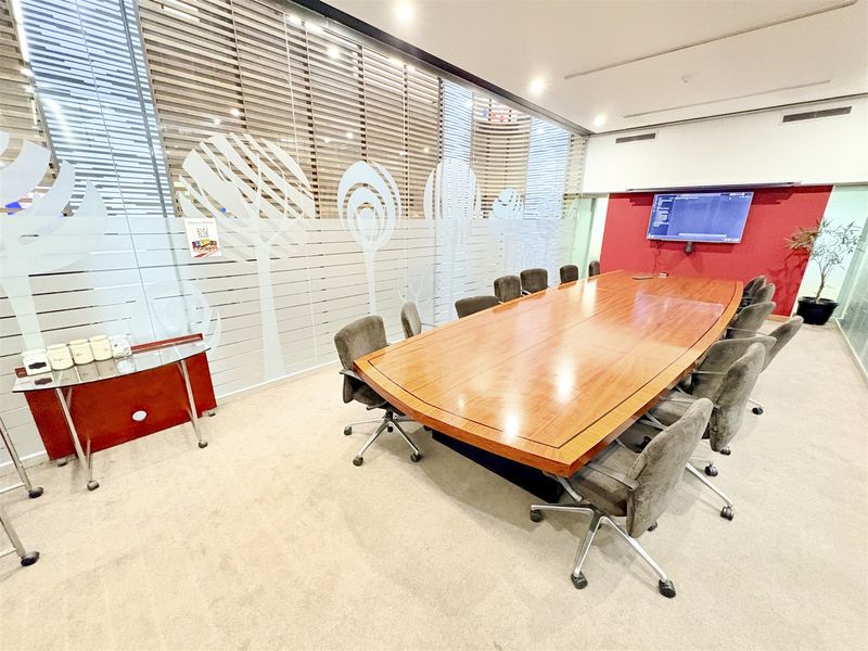A professional office boardroom featuring a large wooden conference table, frosted glass partitions, and a wall-mounted display screen.