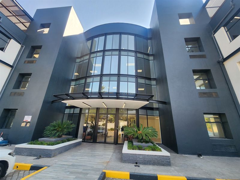 Modern office building entrance featuring a striking glass facade, contemporary dark exterior finish, and a welcoming paved walkway with integrated planters.