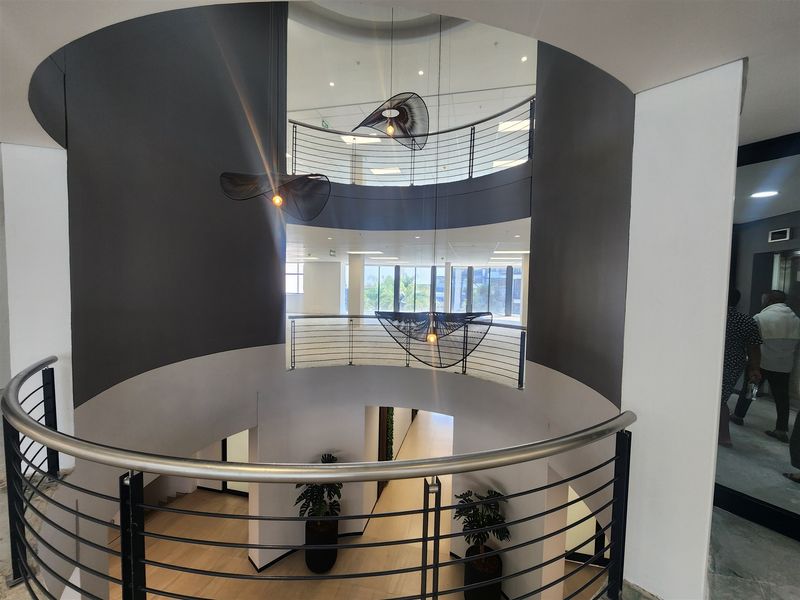 A modern, multi-level office atrium featuring a striking curved staircase, sleek metal balustrades, and contemporary pendant lighting in an open, airy architectural design.