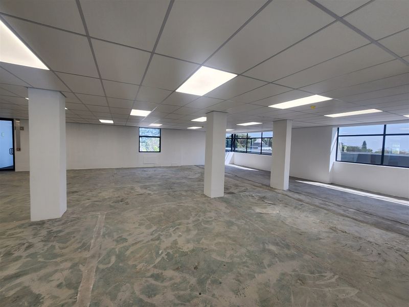 A spacious, open-plan office area featuring a suspended ceiling, recessed lighting, structural columns, and large windows providing ample natural light.